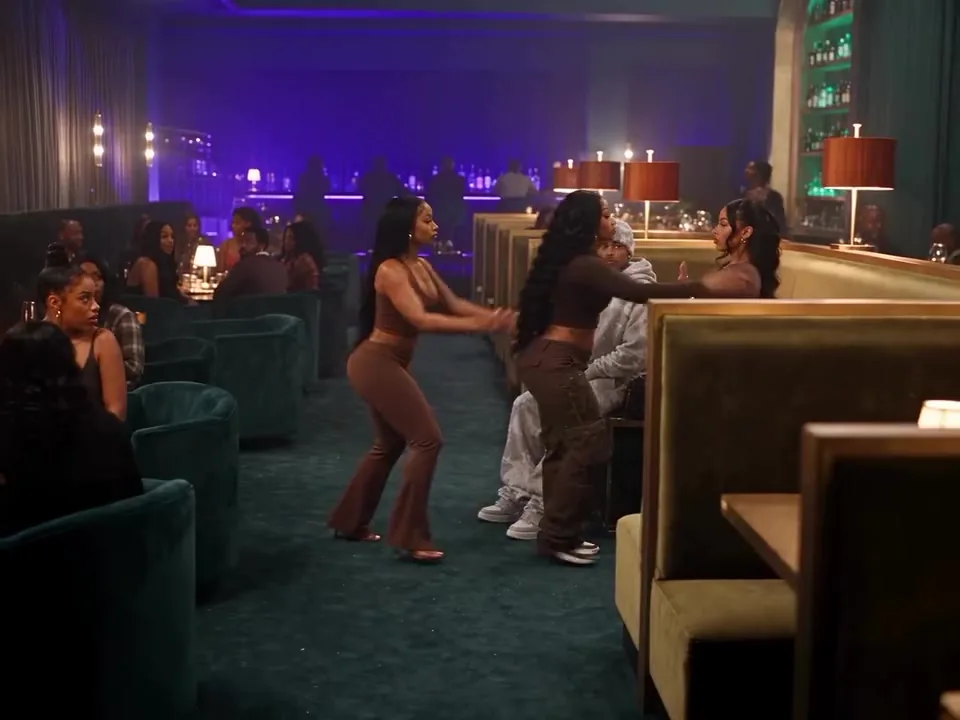Wide shot of a lounge bar with green velvet booths and purple lights as an argument breaks out